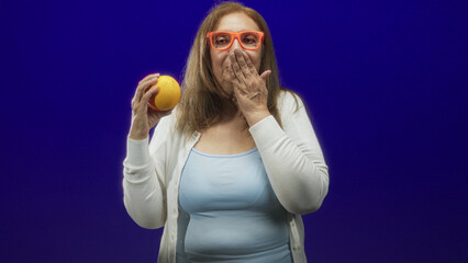 Senior woman holding orange fruit, hand covering mouth and wearing orange glasses in studio;...