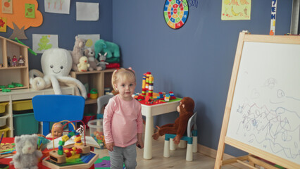 Fototapeta premium Little blonde girl stacks colorful blocks at a plastic play table amid plush toys and shelving in a preschool building; curiosity wonder imagination play.