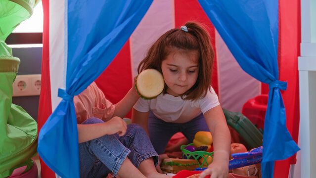 Two children reaching hands for a toy sandwich inside a play tent in building; imaginative friendship playful.