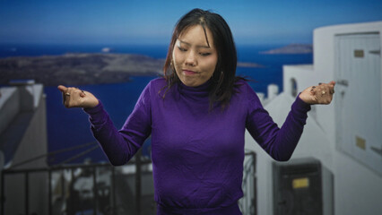 Obraz premium Woman in purple turtleneck raises both hands in a shrug gesture on a white seaside building terrace overlooking blue sea; quiet contemplation.