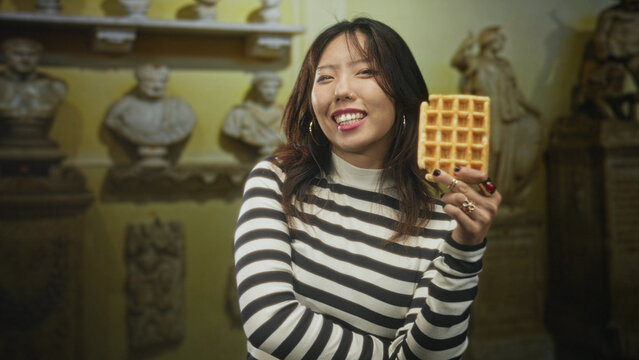 Young chinese woman holding a waffle up near her face, smiling with eyes closed, striped sweater and nose ring, statues visible in building; playful leisure.