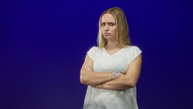 Woman with crossed arms, furrowed brow and wristwatch wearing white blouse in studio blue backdrop; disapproval resolve.