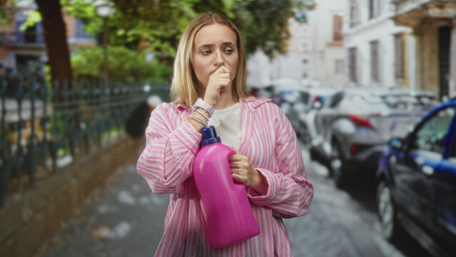 Woman holding pink detergent bottle covers mouth with hand in street while grimacing and clutching the bottle; odor disgust.