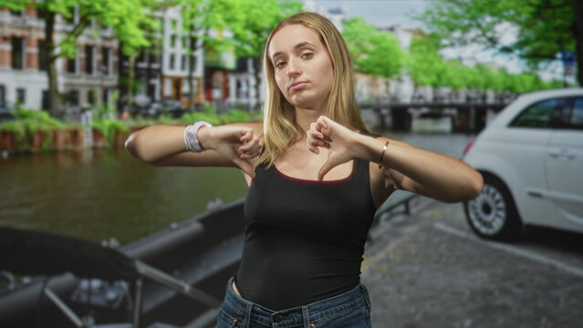 Woman wearing jeans and tanktop showing thumbs down gesture and pouting on a street canal beside a parked car; disapproval rejection attitude.