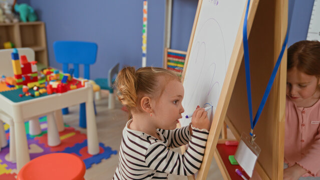 Girl draws and erases on easel with marker and hand in building near toy table, shelf, colorful playmat and blue chair; playful focus.