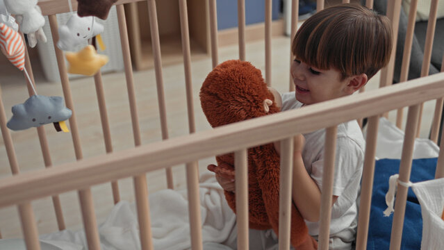 Boy hugging a brown teddy, head resting on the plush while sitting inside a building nursery crib with hanging mobile and blankets; comfort security.