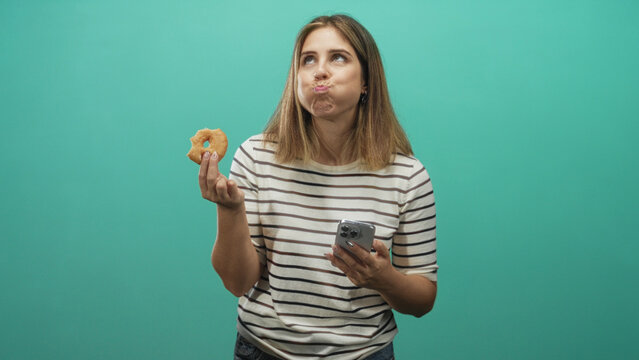 Woman in striped shirt and jeans puffing cheeks while holding a donut and smartphone in a studio; playful curiosity.