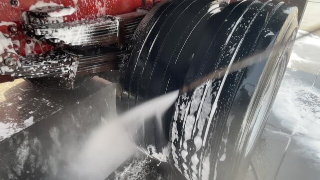 Close view of a wet truck tire being washed with a pressure stream, as white foam and water cover the rubber and wheel area during heavy vehicle cleaning. during detailed truck care process.