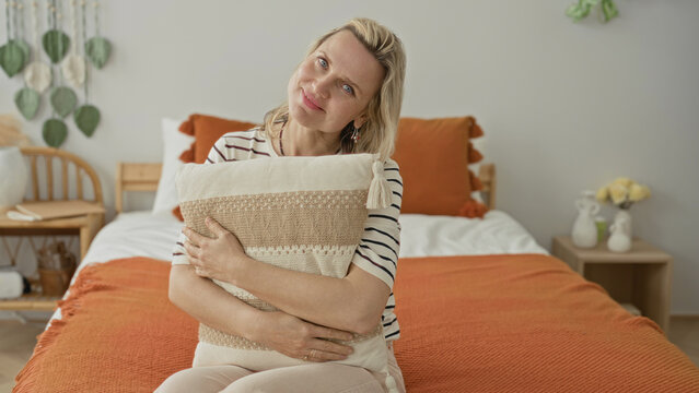 Woman hugging pillow on bed inside a building, hands clasped over cushion and orange throw, seated in gentle posture; quiet contemplation solitude.