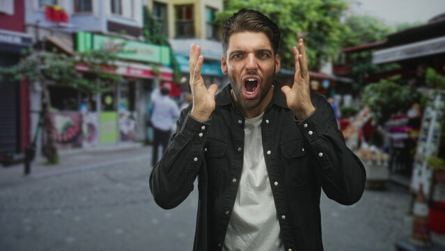 Man with beard, hands to ears, mouth open, wearing black shirt and white tee, standing amid stalls and storefronts in a busy street market scene; surprise urgency.
