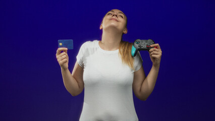 Woman holding a gamepad and a credit card, head tilted back with closed eyes in studio wearing white t shirt  satisfaction. © Krakenimages.com