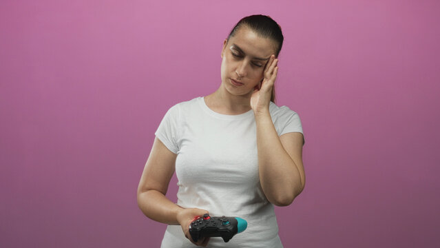 Woman holding a gamepad, pressing temple with fingers and frowning in a pink studio wearing a white tee and ponytail; frustration gaming fatigue.