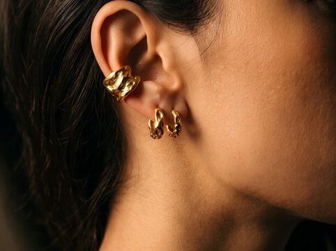 Earrings Collection Featuring Textured Gold Ear Cuff and Huggie Hoops on Woman Ear Close Up in Dramatic Studio Lighting for Luxury Jewelry Fashion Brand Advertising Lookbook and Editorial Promotion