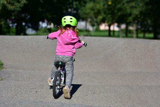 Young girl wearing a safety helmet riding a bicycle on a pump track in a sunny green park, practicing cycling skills and enjoying active outdoor recreation, exercise and childhood adventure.