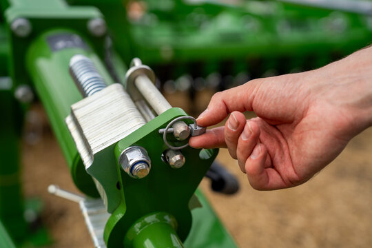 Human hand pulling metal linchpin retainer clip from agricultural machine cylinder mechanism to adjust depth setting on farming equipment outdoors.