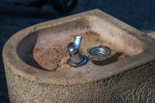 A close-up view shows a weathered stone public drinking fountain with a shiny chrome spout under bright outdoor sunlight.