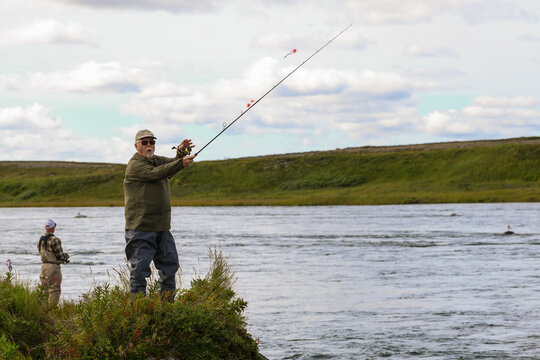 Two anglers cast lines into the Egegik River in Alaska, surrounded by grassy banks and soft clouds, capturing a peaceful outdoor fishing moment.