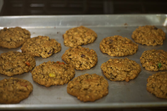Freshly baked oatmeal cookies rest on a metal tray in warm kitchen light, showing texture and golden color.