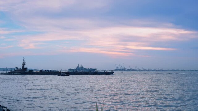 Diverse naval vessels moves by the waterscape slowly. Port infrastructure in the backdrop. U.S. Navy.