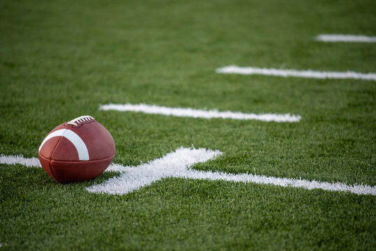 A closeup of an american football resting on the green turf of a stadium field with white yard line markings visible in the background