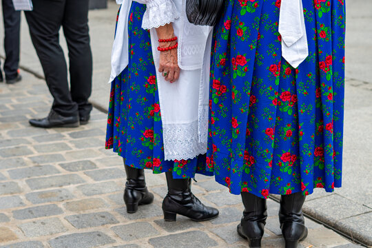 Close-up of the lower part of traditional folk costumes, featuring blue skirts with red rose patterns, white lace aprons, and black leather boots, standing on a paved street, natural daylight.