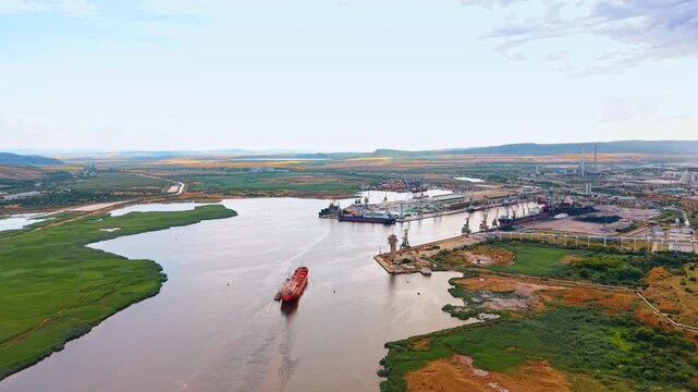 Red cargo ship navigating wide river channel near industrial port. Vessel travels through calm water flanked by green marshland and harbor cranes.
