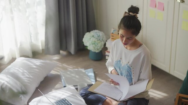 Woman working on laptop sitting on floor by bed, focused studying with sticky notes and books, cozy remote work from home routine in bright bedroom concept