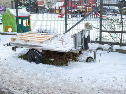 Small metal utility trailer with wooden pallet stands under fresh snow beside a fenced playground. Practical winter transport scene for trailer storage, towing equipment, cargo hauling, maintenance, s