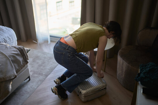 Person Sits on Suitcase in Hotel Room to Close It