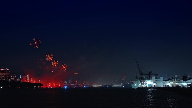 Independence Day fireworks display over water in New York. Red fireworks explode over dark river near industrial harbor and distant city skyline.