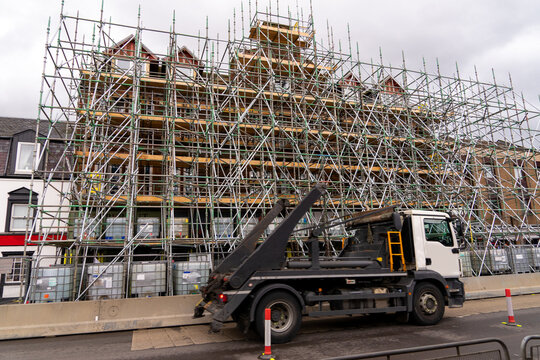 The historic a multi-story  large brick building covered in extensive green metal scaffolding, with wooden walking platforms for renovation work. Commercial rigid chassis skip loader is driving past.