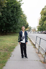 Young Asian male wearing a stylish outfit stands confidently on a sidewalk surrounded by greenery. © VAshowcase