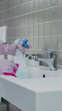 Close up of hands wearing rubber gloves spraying detergent and wiping chrome faucet with cloth