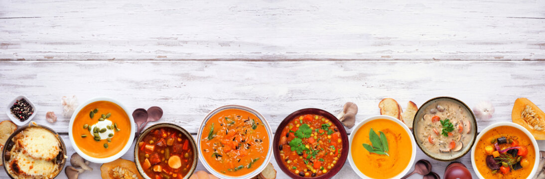 Assortment of delicious homemade warm and cozy autumn soups. Top view bottom border on a white wood banner background. Pumpkin, squash, sweet potato, mushroom, French onion and hearty vegetable.
