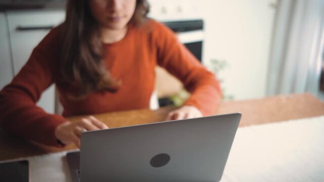 Young Hispanic woman sitting in her home kitchen while opening her laptop, highlighting technology, productivity, digital lifestyle, and everyday domestic life.
