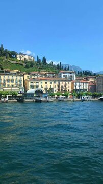 Scenic ferry view of Menaggio waterfront on Lake Como, Italy, with colorful lakeside buildings, green hills and mountains on a sunny summer day