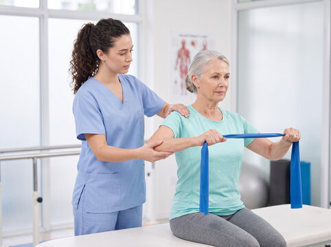 young female physiotherapist guiding senior woman during seated band assisted shoulder flexion rehabilitation in clinic