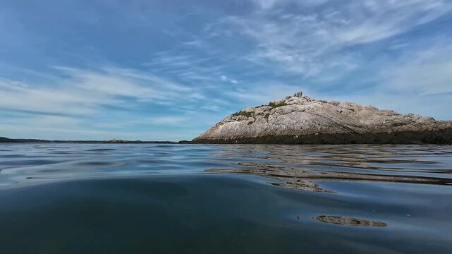 POV camera captures a quiet rocky island rising from rolling blue ocean waters beneath a vast summer sky, creating a peaceful scene of coastal solitude and natural serenity.