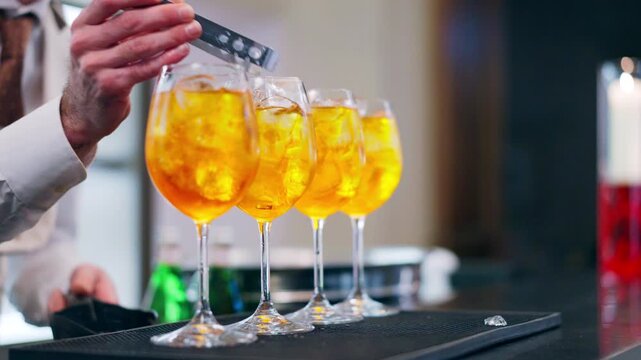 Preparation of traditional Aperol Spritz cocktail at the bar counter, bartender adding ice cubes into the glasses with fizzing drink, refreshing aperitif concept