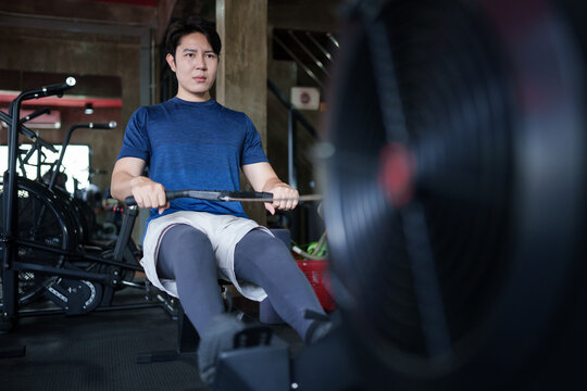 Young Asian man exercising on rowing machine during cardio workout in modern fitness gym