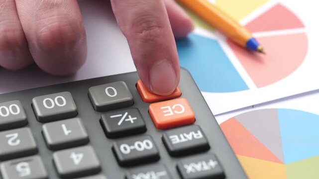 Close-up of a businessman's finger repeatedly pressing the orange ON button on a calculator while analyzing financial pie charts on a desk.