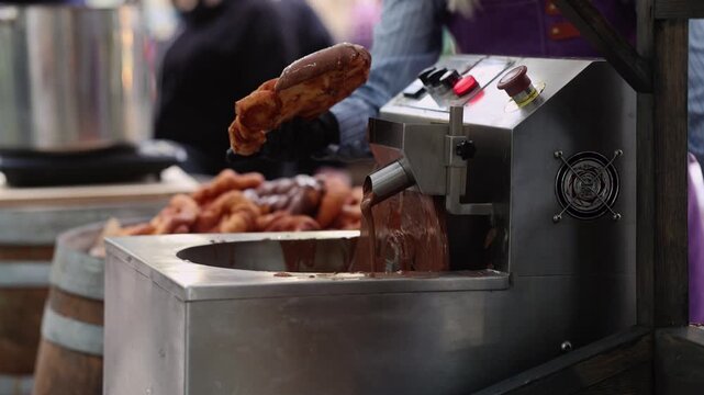 Vendor covering churro with melted chocolate using machine at street food market. Sweet dessert preparation at fair stall. Fresh churros with chocolate sauce, street food cooking concept