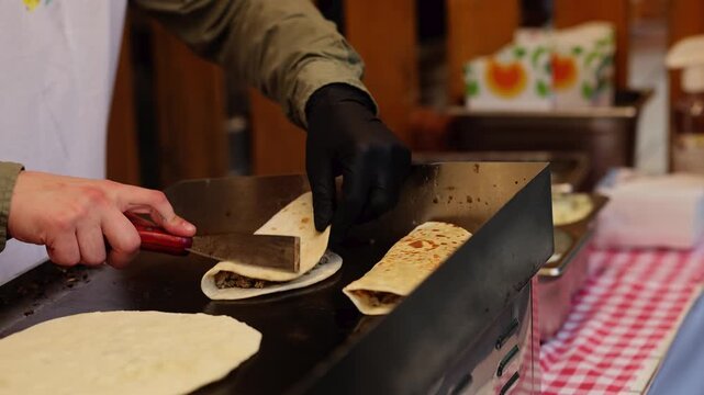 Chef cooking folded tortilla wraps with meat filling on flat grill at street food stall. Cook in black gloves preparing quesadilla with spatula. Fast food preparation concept