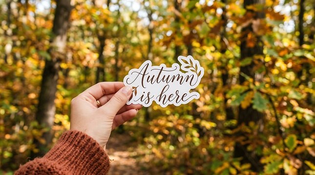 Person holding autumn is here sticker against a blurred background of forest