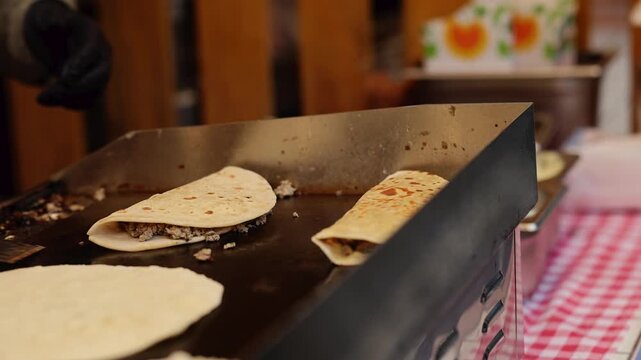 Chef cooking folded tortilla wraps with meat filling on flat grill at street food stall. Cook in black gloves preparing quesadilla with spatula. Fast food preparation concept