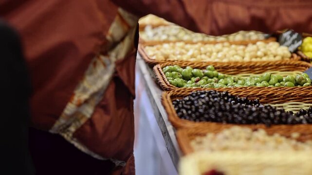 Woman choosing nuts and olives at market stall. Baskets with healthy snacks and dried fruits on counter. Shopping for organic food at street market concept.