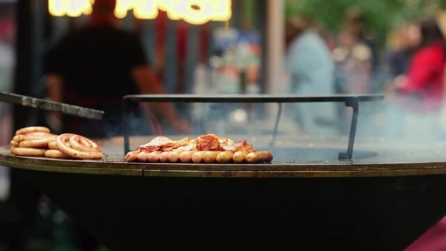 Grilled sausages cooking on round flat grill at street food market. Smoke rising over fried sausages with bacon at outdoor food festival stall