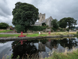 Killaloe Ireland 6th augus 2026. Ancient stone church stands beside calm water. Large tree frames the scene with dense green foliage. Red cranes rest on grassy bank, adding bold color. Still water mir © ANTONIO