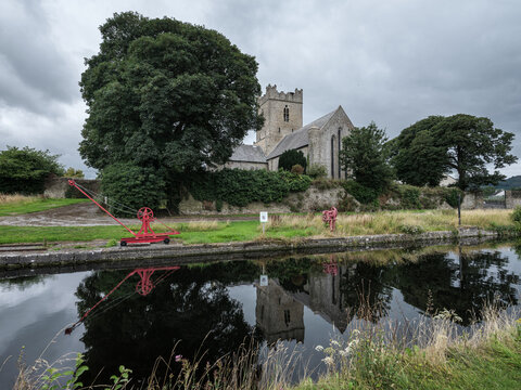 Killaloe Ireland 6th augus 2026. Ancient stone church stands beside calm water. Large tree frames the scene with dense green foliage. Red cranes rest on grassy bank, adding bold color. Still water mir