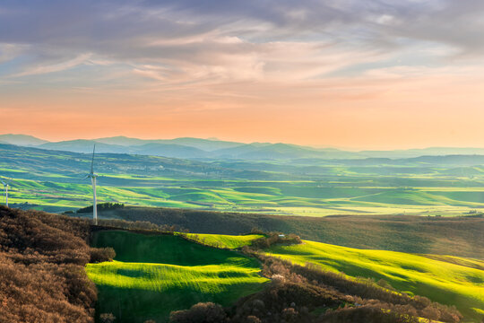 green valley with nice farmland fieds and hills with wind turbines on agricultural landscape. Beautiful rural landscape with cloudy sky on background